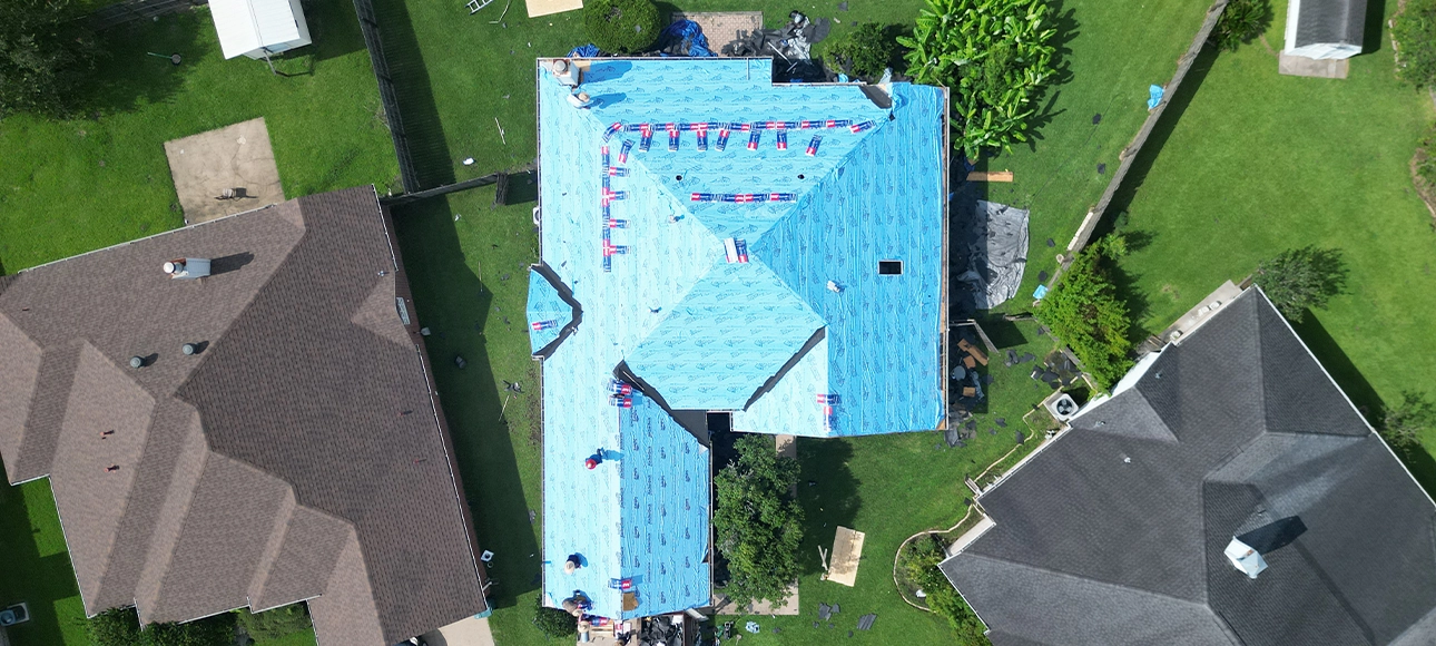 The same roof fully covered in blue synthetic underlayment with shingle bundles staged along the ridges