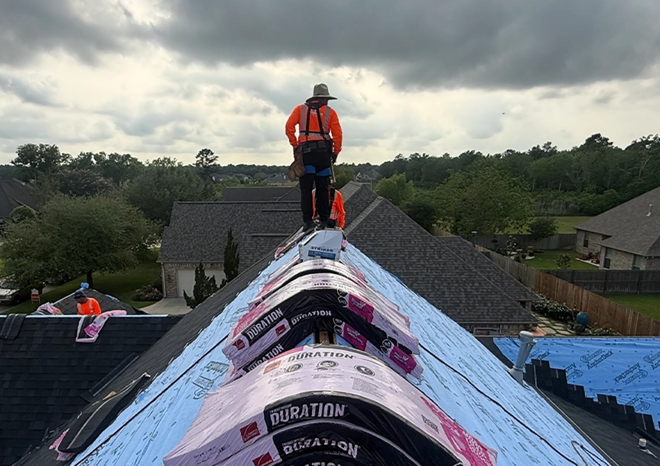 TX Roofing crew member standing on a roof ridge beside bundles of ridge cap shingles under storm clouds
