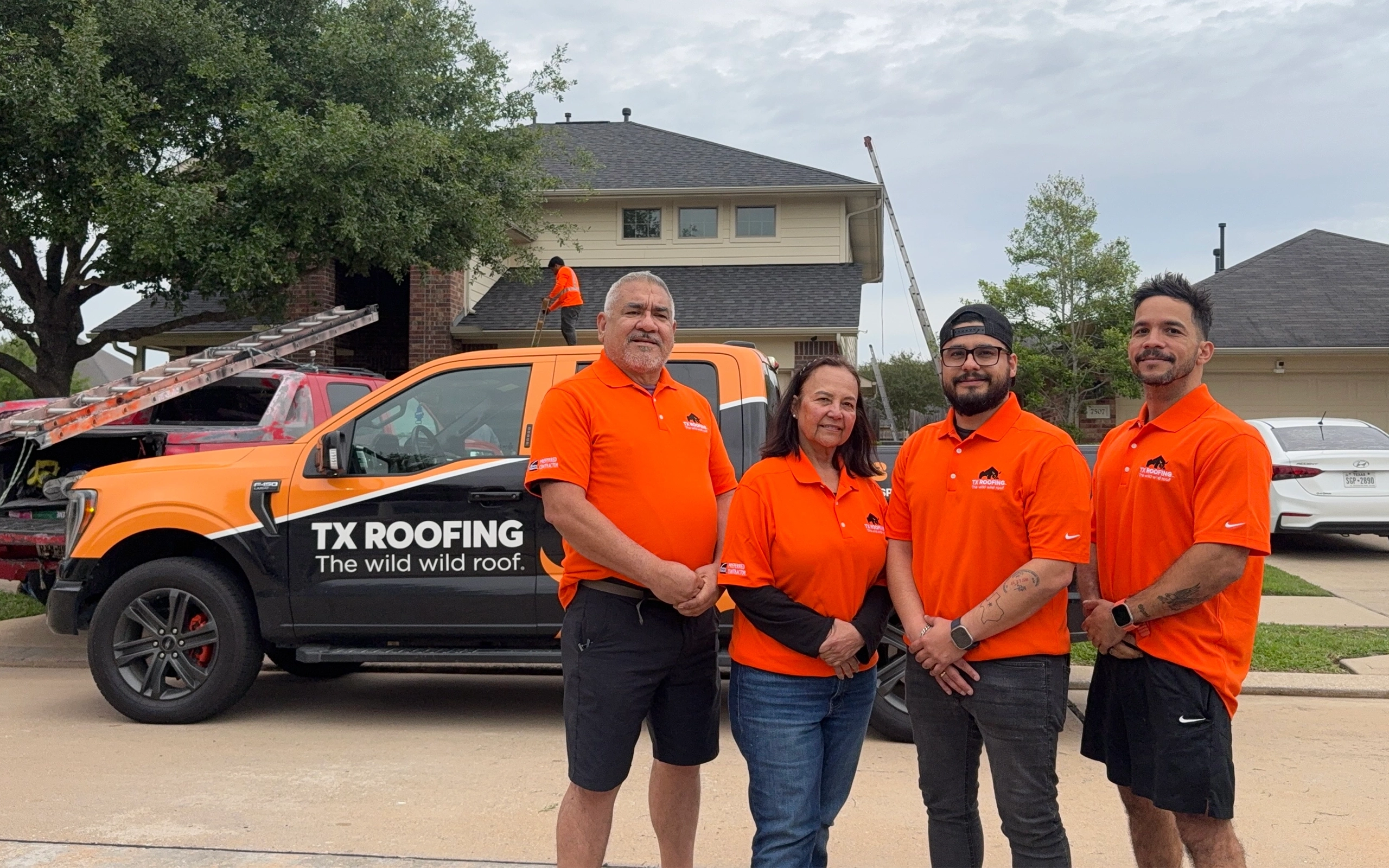 TX Roofing and Services team in orange company polos beside a branded truck while a roofer works on the house behind them